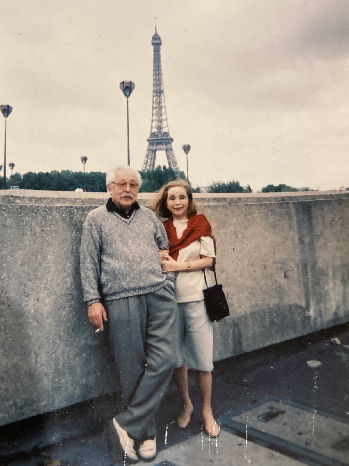 Fotografía antigua de una mujer y un hombre mayor posando junto a un muro de piedra en París, con la torre Eiffel al fondo