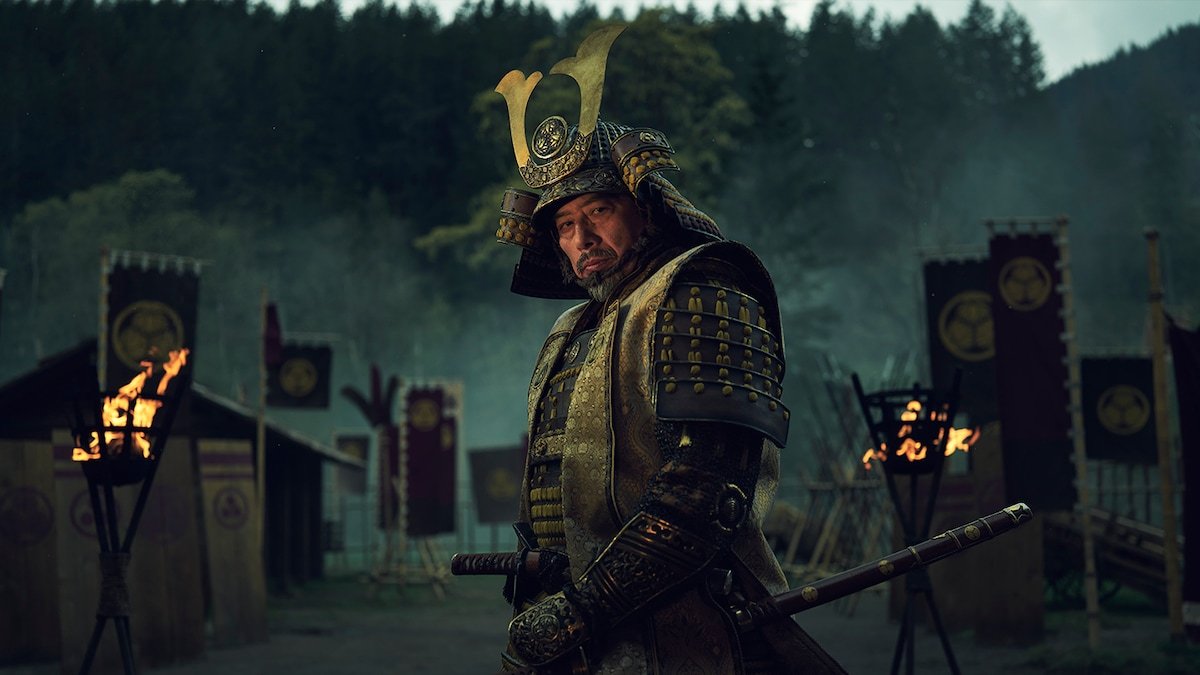 Hiroyuki Sanada con armadura de samurái y casco dorado en un campamento con estandartes y braseros encendidos